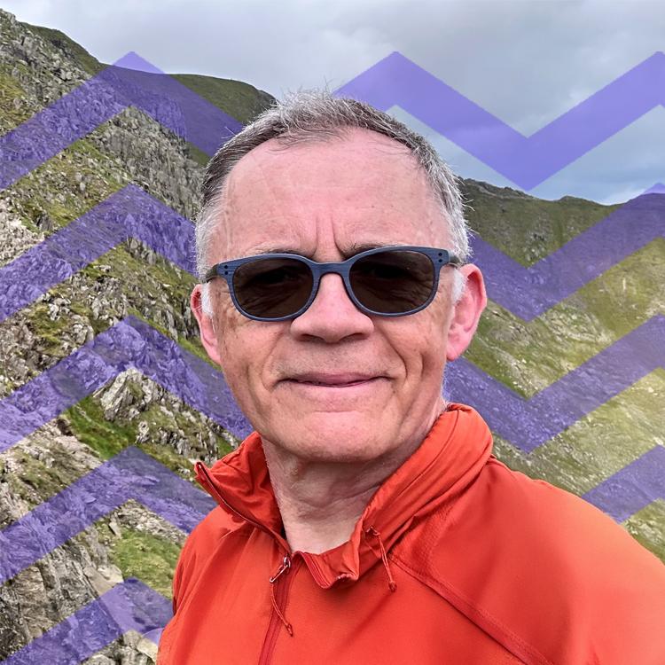 Man wearing sunglasses and an orange jacket standing in front of a hill and a purple mountain pattern