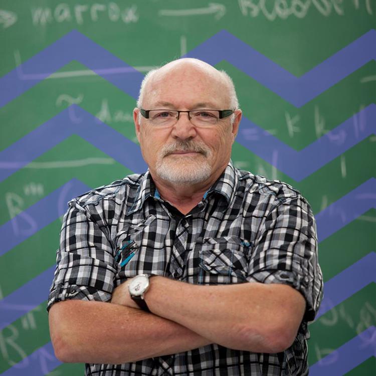 Man wearing glasses and a white beard standing in front of a green blackboard and purple mountain pattern