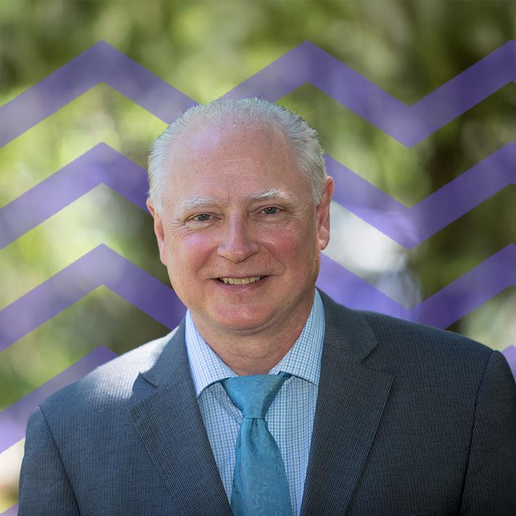 Smiling man wearing a blue suit and blue tie with a purple mountain pattern behind him