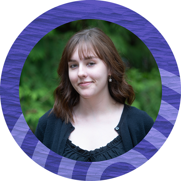 Young woman with brown hair and a dark top and standing front of trees