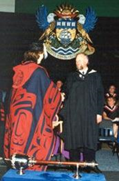 Male student in black gown on stage at graduation ceremony receiving a diploma from a woman in ceremonial First Nations attire